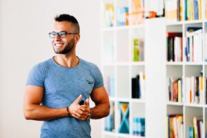 Ein l&auml;chelnder Mann mit Brille und blauem T-Shirt steht in einem hellen Raum vor einem B&uuml;cherregal, das mit bunten B&uuml;chern gef&uuml;llt ist.
