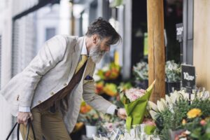 A man in an elegant suit selects flowers in a store. He is holding a bouquet of pink flowers in his left hand. Signs show prices of €10.00 and €20.00.