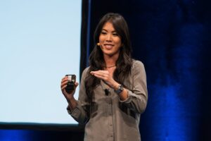 During a presentation, a person holds a small object in their hand and speaks into the microphone. The background is dark and the person is wearing a gray shirt.
