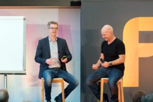 Two men are sitting on bar stools holding microphones. They are smiling and talking. A gray wall with a large yellow letter F can be seen in the background. On the left is an empty flipchart.