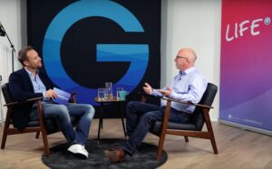Two men are sitting in armchairs and talking. One is holding cards in his hand. Large letters G and LIFE are visible in the background. A jug of water and cups stand on a table between them.