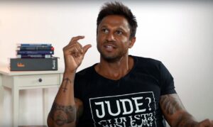 A person is sitting, raising a finger as if explaining something, in a modern, bright room. There is a table with books in the background. Text on the T-shirt: "JUDE? YOU GOT A PROBLEM?".