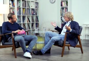 Two men sit opposite each other in armchairs and discuss in a modern room with bookshelves in the background. One of them is holding a book with the title LIFE.
