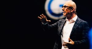 A man with glasses, gesticulating, wearing a checked jacket on a stage with a dark background and a blurred round logo in the background. No other text elements are recognizable.