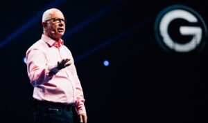 A man with a bald head and glasses wears a pink shirt and speaks while gesticulating. He is standing on a stage while an illuminated G appears in the background.