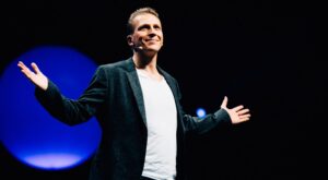 A man stands on a stage with his arms outstretched, wearing a microphone on his ear and smiling. In the background is a large blue circle on a dark background.