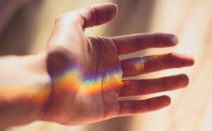 An outstretched hand catches a rainbow light that is projected across the palm. Soft, natural light illuminates the scene in a quiet room.