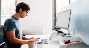 Eine Person mit Brille arbeitet konzentriert an einem Laptop, umgeben von Schreibtischutensilien. Im Hintergrund steht ein gro&szlig;er Monitor in einem hellen, modernen B&uuml;ro mit Fenstern.