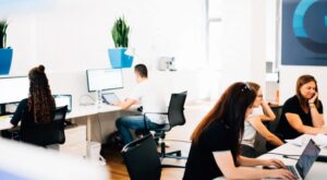 Several people work at computers in a modern office. Some are sitting at desks and concentrating on screens or notes. Green plants and light-colored walls are visible in the background.