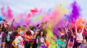 Menschen feiern fr&ouml;hlich, werfen bunte Pulverfarbe in die Luft. Die Menge ist dicht gedr&auml;ngt, lacht und hebt die Arme. Der Himmel ist blau und die Szene wirkt lebendig und euphorisch.