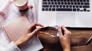 One hand holds a partially opened chocolate bar in front of a laptop on a wooden table. A pink cup and a notebook are to the left.