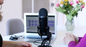 A microphone is placed on a table and is apparently being used for a recording, while an open laptop and a bouquet of flowers on a white table can be seen in the background.