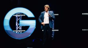 A person in a blue suit speaks on a stage and gesticulates. Behind her stands a large, illuminated G in front of a dark background with horizontal light bars.