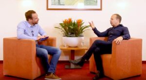 Two men sit in orange armchairs and talk in a bright room. A table with a large plant stands between them. A picture hangs on the wall in the background.