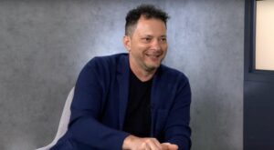 Ein l&auml;chelnder Mann in einem blauen Sakko gestikuliert mit der rechten Hand. Er sitzt vor einer grauen Wand, die eine ruhige, neutrale Umgebung schafft.