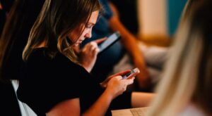 A person is holding a smartphone and concentrating on it in a room with other seated people who are also busy with their mobile devices. The atmosphere seems concentrated and shielded.