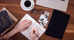 A person is writing in an open notebook on a wooden table. Next to it are a pair of glasses, a tablet, a laptop, a pile of books, a coffee and Polaroid photos.
