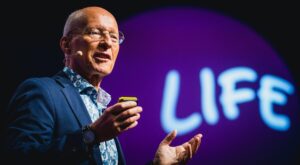 Ein Mann tr&auml;gt ein Headset, h&auml;lt einen kleinen Gegenstand und spricht leidenschaftlich auf einer B&uuml;hne. Im Hintergrund befindet sich ein violetter Kreis mit dem gro&szlig;en wei&szlig;en Text LIFE.