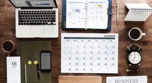 Ein aufger&auml;umter Schreibtisch zeigt einen Laptop, offene Notizb&uuml;cher, Kalender, eine Kaffeetasse, einen Wecker und ein Smartphone. Alles liegt ordentlich auf einer Holzoberfl&auml;che, arrangiert f&uuml;r produktive Arbeit.