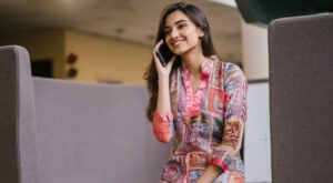 A person wears a colorful, patterned dress, smiles while talking on the phone and sits between gray partitions. The background shows an indoor environment with soft lighting.
