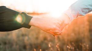 Two hands hold each other tenderly in a warm sunset, surrounded by a blurred, golden field background that conveys a peaceful and intimate mood.