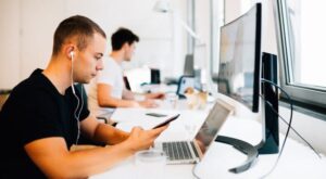 A man with headphones is sitting at a desk, using a smartphone next to a laptop. In the background, another person is working on a computer. The environment is an office.