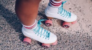 Wei&szlig;e Rollschuhe mit blauen Schn&uuml;rsenkeln gleiten &uuml;ber eine grobe, gemusterte Oberfl&auml;che. Die Rollen sind rosa, und der Tr&auml;ger tr&auml;gt Socken mit roten Streifen. Der Hintergrund zeigt einen Wechsel zwischen Beton und Sand.
