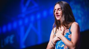 A woman with long hair wears a blue dress with a floral pattern. She holds her hands on her chest, eyes closed, possibly speaking, with a microphone to her ear, in the spotlight on a stage. In the background, shadowy blue letters.