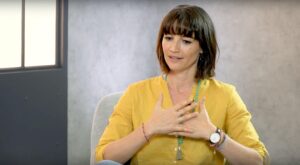 A woman in a yellow blouse sits on an armchair, gesticulating with her hands in front of her chest. The background shows a gray wall and a window with a dark frame.