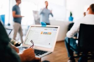 Ein Laptop mit ge&ouml;ffnetem Browser zeigt eine Website. Eine Person schreibt Notizen auf Papier. Im Hintergrund steht jemand und spricht, w&auml;hrend andere sitzend zuh&ouml;ren. Helles B&uuml;roambiente.