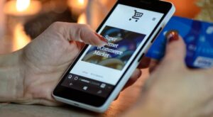 A smartphone is held with one hand, the screen reads "Super Internet eCommerce Market". Another hand holds a blue credit card. Lights shine in the background.