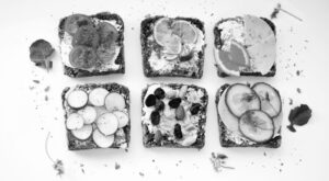 Sechs belegte Brotscheiben in einer 2x3-Anordnung, jede mit unterschiedlichen Bel&auml;gen wie Tomaten, Zitrone, Radieschen, N&uuml;ssen, Trockenfr&uuml;chten, Gurken und K&auml;se. Umgeben von verstreuten Kr&auml;utern und Bl&auml;ttern.