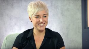 A person with short, blond hair smiles in a relaxed posture. She wears a black shirt and sits on a gray chair in front of a neutral gray background.