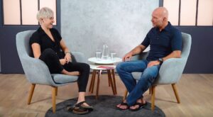Two people sit opposite each other in gray armchairs and talk in a modern, light-flooded environment. A small table with glasses and a bottle of water stands between them.
