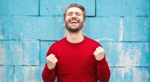 A man in a red sweater laughs joyfully with his fists clenched. He is standing in front of a blue brick wall, which forms a contrasting background.