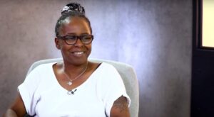 A person with glasses and a white top smiles while sitting on a gray chair. The background is plain gray, creating a relaxed, clear conversational atmosphere.