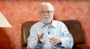 A man with a white beard and glasses sits on a brown leather armchair, gesticulating with his left hand. He is wearing a light blue shirt. The background is orange with a textured surface.