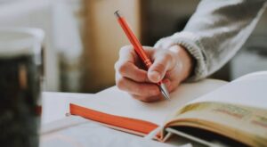 A hand holds a red pen and writes in an open notebook. The background shows a cup out of focus, indicating a cozy environment. There is no text.