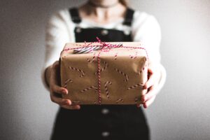 A person in black overalls and a white top holds out a Christmas-wrapped gift decorated with candy canes and red string. Blurred, neutral background.