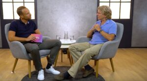 Two men sit in gray armchairs and talk. One of them is holding a magenta pad with the word LIFE written on it. They are in a room with a wooden floor and modern design accents.