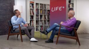 Two men sit opposite each other in armchairs and talk in a modern room with bookshelves in the background. A red poster with the word "LIFE" is visible on the wall.