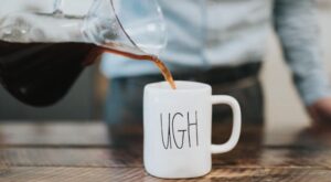 Eine wei&szlig;e Tasse mit dem Text UGH wird mit Kaffee aus einer Glaskaraffe gef&uuml;llt. Der Hintergrund zeigt eine verschwommene Person in einem Hemd hinter einem Holztisch.