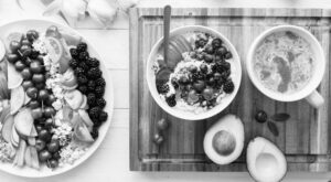 A breakfast tray presents a bowl of muesli, fresh berries and nuts, with a cup of coffee next to it. On the left is a full salad plate with vegetables and fruit. A healthy meal.