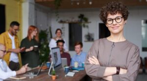 Eine Person steht l&auml;chelnd mit verschr&auml;nkten Armen im Vordergrund, w&auml;hrend im Hintergrund mehrere Personen in einem modernen B&uuml;ro besch&auml;ftigt sind, um einen Tisch arbeiten und sich unterhalten.