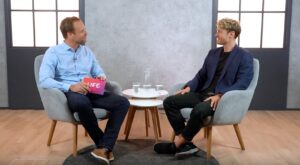 Two men are sitting in gray armchairs, smiling and talking. One man is holding a card labeled LIFE. Between them is a table with glasses of water. The room has a wooden floor and a gray wall.