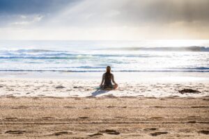 Eine Person sitzt ruhig im Lotossitz am Sandstrand, blickt aufs Meer, w&auml;hrend die Sonne durch die Wolken hindurchstrahlt. Der Ozean wirkt friedlich mit sanften Wellen.
