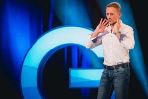 A man in a white shirt and blue jeans speaks gesticulating in front of a large, glowing letter G. He is on a stage surrounded by blue lights.