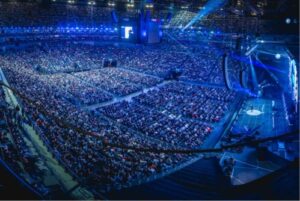 A large crowd sits in a brightly lit stadium, while blue lights illuminate the stage right. Screens broadcast the event to spectators further away.
