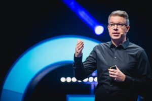 A person is wearing a microphone and giving a lecture, gesticulating with his right hand and holding a small object in his left. The background is dark with glowing blue circles.