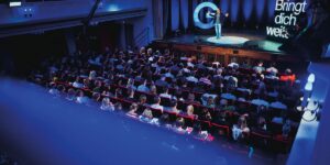 Ein Redner steht auf einer B&uuml;hne und spricht vor einem gro&szlig;en Publikum in einem Theatersaal. Rechts neben ihm leuchten die Worte: &bdquo;Bringt dich weiter&ldquo; in heller Schrift auf.